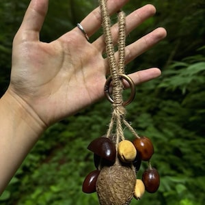 May include: A handmade keychain featuring a cluster of natural seeds and nuts in various shades of brown and tan. The seeds are attached to a woven jute cord, with a metal ring at the top for easy attachment to keys or a bag.