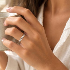 May include: A slim, polished silver-toned band ring featuring engraved black text that reads 39.9039° N 89.7541° N. The ring has a smooth, flat surface and a minimalist design, encircling the finger with clear, legible geographic coordinates.
