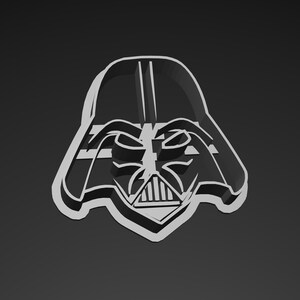 May include: A white, three-dimensional cookie cutter in the shape of Darth Vader's helmet. The design features the iconic mask with its distinct features, including the mouth grill and eye slits, set against a dark gray background.