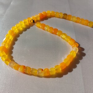Op de afbeelding: Een ketting bestaande uit kleine, vierkant gesneden kralen in tinten geel en oranje. De kralen zijn aan elkaar geregen tot een doorlopende lus, met een goudkleurige sluiting. De ketting wordt getoond op een witte, getextureerde ondergrond.