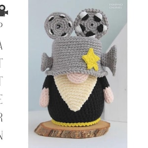 Könnte beinhalten: Gehäkelte Zwergenpuppe im Filmthema. Die Puppe hat einen grauen Hut mit Filmrollen-Details, einen gelben Stern und ein schwarz-cremefarbenes Outfit. Die Puppe steht auf einem Holzsockel. Das Wort "PATTERN" ist auf der linken Seite des Bildes.