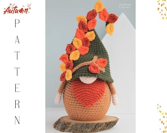 Op de afbeelding: Een handgemaakte gehaakte kabouter met een bruin lichaam, een groene hoed versierd met herfstbladeren en een rode hartvormige baard. De kabouter wordt gepresenteerd op een houten schijf, met de woorden "Autumn" en "Pattern" in de afbeelding.