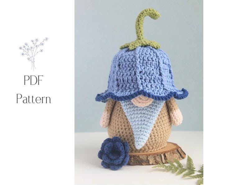 May include: A crocheted gnome doll with a blue bell-shaped hat and a light blue beard. The gnome has a tan body, pink hands, and a small blue flower. The words "PDF Pattern" are in the image.