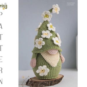 May include: A handmade crochet gnome with a green body and hat, adorned with white and yellow flower accents. The gnome has a beige beard, pink hands, and is displayed on a wooden slice. The image includes the text "Spring Pattern".