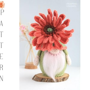 Op de afbeelding: Een handgemaakte gehaakte kabouter met een grote oranje bloemenhoofdtooi. De kabouter heeft een groen lichaam, een roze en witte baard en wordt gepresenteerd op een houten schijf. Het woord "PATTERN" staat verticaal links uitgelijnd.