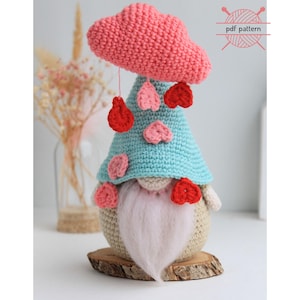 May include: A handmade crochet gnome with a light blue hat adorned with pink and red hearts, topped with a pink cloud. The gnome has a beige body, a fluffy white beard, and sits on a wooden slice. The image includes the text "pdf pattern".
