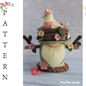 May include: A handmade crochet gnome with a bird in a nest design. The gnome has a long white beard, a green body, and a brown nest with a pink and cream-colored bird. The design includes pink and yellow flowers and the word "PATTERN".