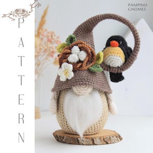 Puede incluir: Un gnomo de ganchillo hecho a mano con una larga barba blanca, que lleva un sombrero marr&oacute;n adornado con un nido, flores y hojas. Un peque&ntilde;o p&aacute;jaro con plumaje amarillo, negro y naranja se posa en un lazo sobre el gnomo. El gnomo est&aacute; sobre una base de madera.