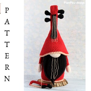 May include: A handmade red and black crochet gnome shaped like a violin. The gnome has a long black beard, a red hat, and white accents. A small wooden bow rests in front of the gnome.
