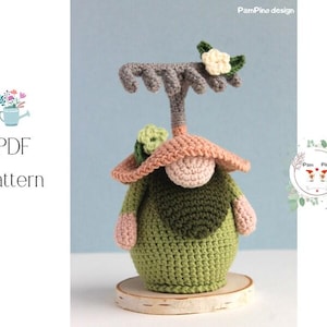 Puede incluir: Una figura de gnomo de ganchillo hecha a mano con un rastrillo, que lleva un atuendo verde, un sombrero color melocotón y una cara beige. El gnomo está sobre una base de madera. La imagen incluye el texto "PDF Pattern".