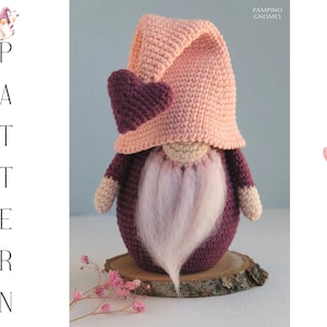 Könnte beinhalten: Ein handgehäkelter Zwerg mit rosa Mütze und herzförmigem Akzent. Der Zwerg hat einen weißen Bart und steht auf einer Holzscheibe. Das Bild enthält das Wort „PATTERN“ und den Text „PAMPINO GNOMES“.