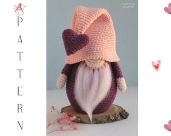 May include: A handmade crochet gnome with a pink hat and a heart-shaped accent. The gnome has a white beard and is set on a wooden slice. The image includes the word "PATTERN" and the text "PAMPINO GNOMES".