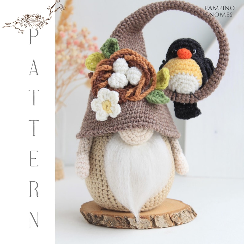 Puede incluir: Un gnomo tejido a crochet con un sombrero marr&oacute;n, barba blanca y un p&aacute;jaro posado en un lazo. El gnomo tiene un nido con huevos y una flor en su sombrero. El p&aacute;jaro es negro, amarillo y naranja. El gnomo est&aacute; sobre una base de madera.