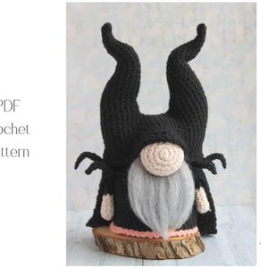 以下が含まれることがあります： 黒いかぎ針編みのノーム。高く湾曲した角、ベージュの鼻、長い灰色のひげがあります。ノームは小さな尖った袖が付いた黒いマントを着ています。ベースは木の輪切りで、「PDF Crochet Pattern」というテキストが表示されています。