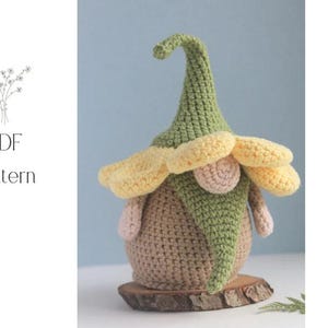 May include: A handmade crochet gnome with a green pointed hat and yellow flower-like accents. The gnome has a tan body, pink nose and arms, and sits on a wooden base. The image also includes the text "PDF Pattern".