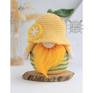 May include: A handmade crochet gnome with a yellow hat, beard, and body, and a green and yellow striped base. The gnome has a lemon slice applique on its hat and is displayed on a wooden slice. The image also includes the text "PATTERN".