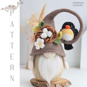 Puede incluir: Un gnomo de ganchillo con un sombrero marr&oacute;n, barba blanca y un p&aacute;jaro en un asa circular. El sombrero del gnomo tiene un nido con huevos y una flor. El texto "PAMPINO GNOMES" es visible.
