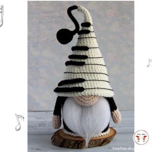 May include: A handmade crochet gnome with a piano key-patterned hat and a long white beard. The gnome's hat is cream-colored with black keys, and it has a black treble clef accent. The gnome is standing on a wooden base.