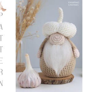 Puede incluir: Una muñeca gnomo de ganchillo con barba y sombrero blancos, que se asemeja a un diente de ajo. La muñeca es de color beige y crema, con un cuerpo redondo y una base de madera. Un bulbo de ajo real está al lado de la muñeca. La palabra "PATTERN" está escrita verticalmente.