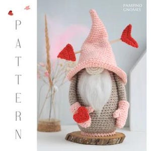 Pode incluir: Um gnomo de crochê feito à mão com um chapéu rosa, corpo cinza e barba branca. O gnomo segura um coração vermelho e tem uma base de madeira. Uma flecha decorativa com penas em forma de coração vermelho está presa ao chapéu. A palavra "PATTERN" é impressa verticalmente à esquerda.