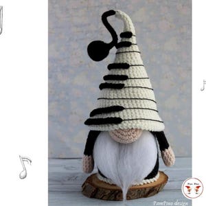 Può includere: Uno gnomo all'uncinetto fatto a mano con un design a tasti di pianoforte sul cappello. Lo gnomo ha una barba bianca, un naso beige e dettagli neri e bianchi. È esposto su una fetta di legno. La parola "PATTERN" è scritta sul lato sinistro dell'immagine.