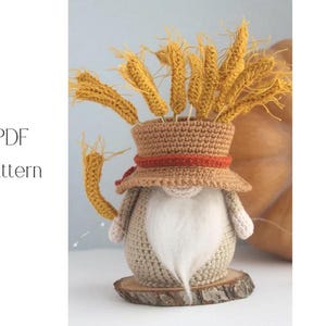 May include: A crocheted gnome figurine with a wheat-like arrangement in its hat. The gnome has a white beard, a brown hat with an orange band, and is set on a wooden base. The text "PDF Pattern" is visible.