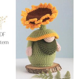 May include: A crocheted gnome figurine with a sunflower head. The gnome is green with a yellow and orange sunflower, a tan nose, and pink arms. The figurine sits on a wooden slice. The words "PDF Pattern" are in the image.