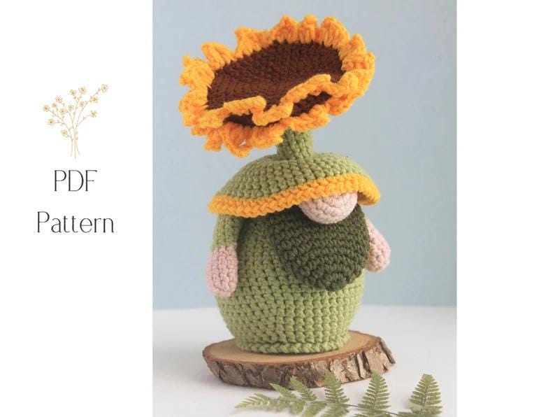 May include: A crocheted gnome figurine with a sunflower head. The gnome is green with a yellow and orange sunflower, a tan nose, and pink arms. The figurine sits on a wooden slice. The words "PDF Pattern" are in the image.