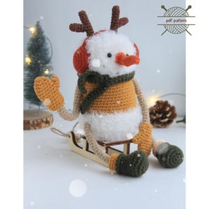 May include: A handmade crochet snowman with reindeer antlers, earmuffs, and a scarf, sitting on a wooden sled. The snowman has a white body, orange and yellow accents, and green mittens and boots. The image includes the word "PATTERN".