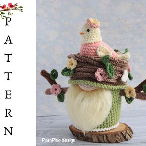 May include: A handcrafted crochet gnome with a bird on a nest. The gnome features a long, fluffy white beard, a green body, and a brown hat decorated with flowers. The bird is pink and cream, with a small yellow beak. The gnome is displayed on a wooden base.