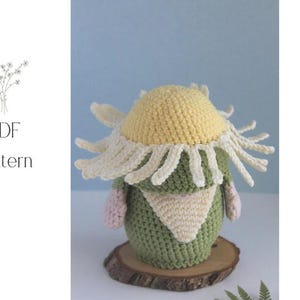 May include: A crocheted gnome doll with a yellow and white flower hat, green body, and a cream-colored scarf. The doll is sitting on a wooden slice. The words "PDF Pattern" are in the upper left corner.