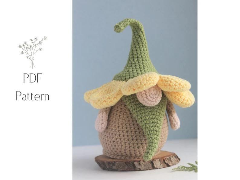 May include: A handmade crochet gnome doll with a green hat, yellow flower-like brim, and a tan body. The gnome is sitting on a small wooden base. The image also includes the text "PDF Pattern".