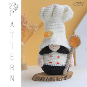 Könnte beinhalten: Ein handgefertigter gehäkelter Kochgnom mit weißer Kochmütze und Uniform, der einen Holzlöffel hält. Der Gnom hat einen schwarzen Schnurrbart und steht auf einer Holzscheibe. Der Text "PATTERN" und "PAMPINO GNOMES" ist enthalten.