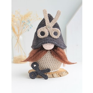 Puede incluir: Un gnomo de crochet que lleva un gorro gris con un par de tijeras en la parte delantera. El gnomo tiene una barba marrón y sostiene un par de tijeras en su mano.