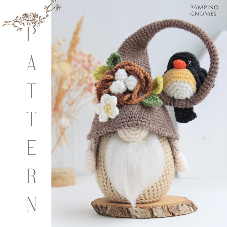 Puede incluir: Un gnomo de ganchillo con una larga barba blanca, que lleva un sombrero marr&oacute;n y sostiene un p&aacute;jaro posado en un lazo marr&oacute;n. El gnomo est&aacute; decorado con un nido, flores y hojas. El texto "PAMPINO GNOMES" es visible.