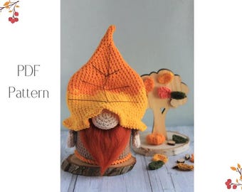 May include: A handmade crochet gnome with a leaf-shaped hat in shades of orange and yellow, a beige face, and a red beard. The gnome sits on a wooden base. The image also includes a wooden tree with decorative leaves and pumpkins. Text on the left says "PDF Pattern".