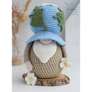 Puede incluir: Muñeca de gnomo de ganchillo con una cabeza azul y verde en forma de globo, una barba blanca y un cuerpo marrón. La muñeca está adornada con un sombrero azul y una flor blanca. La muñeca está sobre una rodaja de madera.