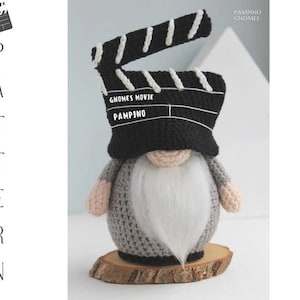 Könnte beinhalten: Handgefertigte Zwergenfigur mit einem Filmklapptafelhut. Der Zwerg hat einen langen weißen Bart, einen grauen Körper und steht auf einem Holzsockel. Der Klapptafelhut ist schwarz mit weißen Akzenten und dem Text "GNOMES MOVIE PAMPINO".