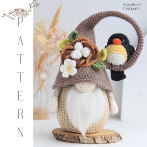Puede incluir: Una mu&ntilde;eca de gnomo hecha a mano con un gorro puntiagudo marr&oacute;n, una barba blanca y un p&aacute;jaro posado en un marco circular. El sombrero del gnomo est&aacute; adornado con un nido, flores y hojas. La mu&ntilde;eca est&aacute; sobre una base de madera. El texto "PAMPINO GNOMES" es visible.