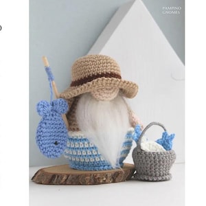 Può includere: Figurina di gnomo all'uncinetto con un cappello beige con una fascia marrone, una lunga barba bianca e pantaloni a righe blu e bianche. Lo gnomo tiene una canna da pesca con un pesce blu e un cestino grigio con pesci blu. La parola "PATTERN" è scritta verticalmente a sinistra.