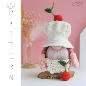 May include: A handmade crochet gnome wearing a chef's hat and apron, topped with a red cherry. The gnome is pink, white, and beige, standing on a wooden slice. The word "PATTERN" is vertically displayed on the left side of the image.