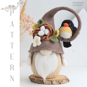 Puede incluir: Una mu&ntilde;eca de gnomo de ganchillo hecha a mano con una larga barba blanca, que lleva un sombrero marr&oacute;n adornado con un nido, huevos y una flor blanca. Un p&aacute;jaro negro y amarillo se posa en un arco marr&oacute;n. El gnomo est&aacute; sobre una base de madera.