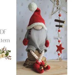 May include: A handmade crochet gnome doll with a red hat, grey sweater, and long white beard. The gnome has red mittens and shoes with bells. The doll is sitting on a wooden surface. A decorative star and bead garland is in the background.