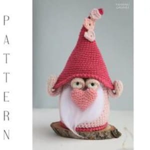 Könnte beinhalten: Ein handgefertigter Häkelzwerg mit rosa Hut und Bart. Der Zwerg hat eine herzförmige Nase und sitzt auf einem Holzsockel. Das Wort "PATTERN" steht vertikal auf der linken Seite des Bildes.