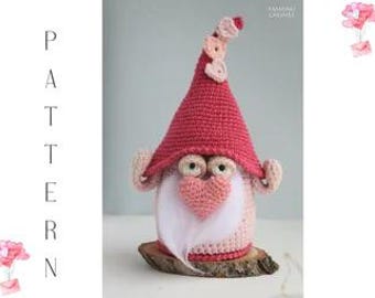Könnte beinhalten: Ein handgefertigter Häkelzwerg mit rosa Hut und Bart. Der Zwerg hat eine herzförmige Nase und sitzt auf einem Holzsockel. Das Wort "PATTERN" steht vertikal auf der linken Seite des Bildes.