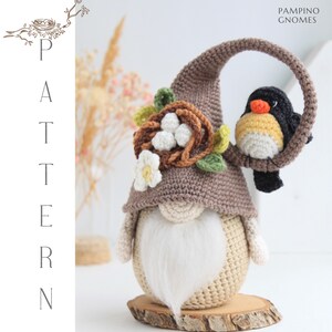 Puede incluir: Una mu&ntilde;eca de gnomo artesanal con una larga barba blanca, que lleva un sombrero marr&oacute;n tejido adornado con un nido y un pajarito en una rama. El gnomo est&aacute; sobre una base de madera. El texto "PAMPINO GNOMES" es visible.