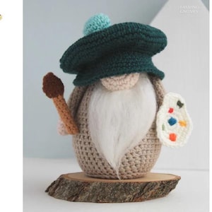 May include: A crocheted gnome artist figurine with a green beret and a white beard. The gnome holds a paintbrush and a palette with colorful paint splotches. The figurine is displayed on a wooden slice.