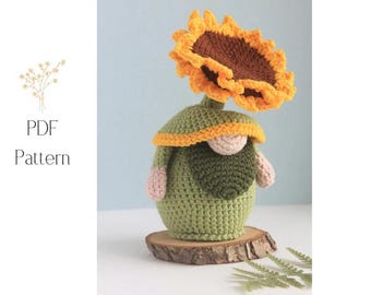 May include: A crocheted gnome with a sunflower head. The gnome is green with a yellow and orange brim, a beige face, and pink arms. It sits on a small wooden slice. The text "PDF Pattern" is on the left.