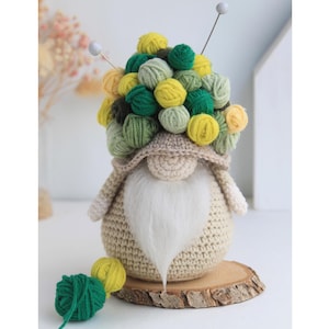 Puede incluir: Un gnomo de crochet con barba blanca y un sombrero hecho de bolas de lana verde y amarilla. El gnomo está sentado sobre una base de madera.