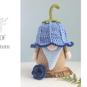 Könnte beinhalten: Eine gehäkelte Zwergenpuppe mit blauem Blumenhut und passender blauer Blume. Der Zwerg hat einen beigefarbenen Körper, einen hellblauen Bart und rosa Hände. Der Text "PDF Pattern" ist sichtbar.